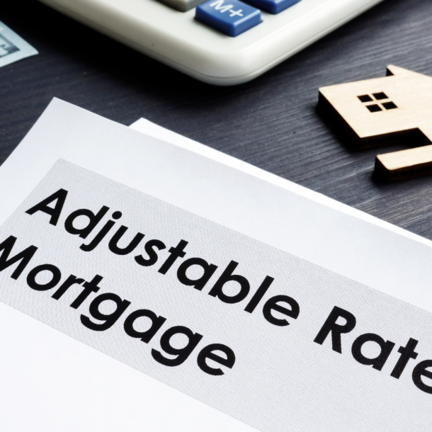 Paper with text that reads "Adjustable Rate Mortgage" from a local mortgage lender sits on a desk with a partial image of a calculator above and a cardboard cut-out of a home to the right.