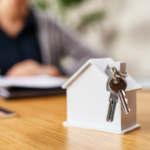 A scale model of a home sits on top of a light wood desk with keys attached to the chimney in the foreground with a customer in the background closing on their new home from the best local mortgage lenders.