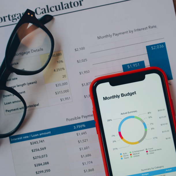 A customer calculates their budget from a trusted local lender on a smartphone sitting on top of a paper that reads "Mortgage Calculator" in the background. A pair of black reading glasses sits on top of the paper.